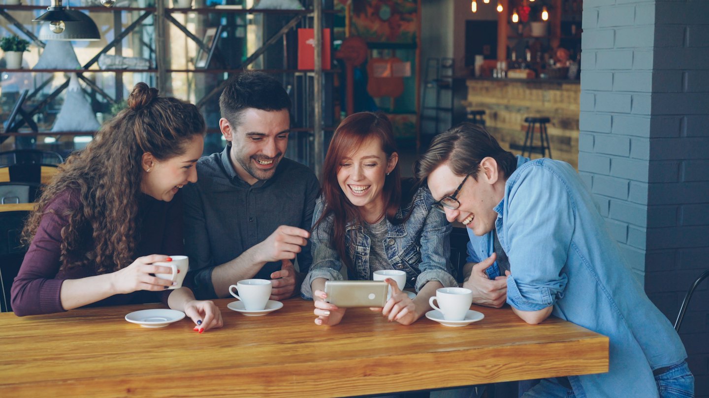Vier Freunde lachen über ein Smartphone an einem Cafétisch.